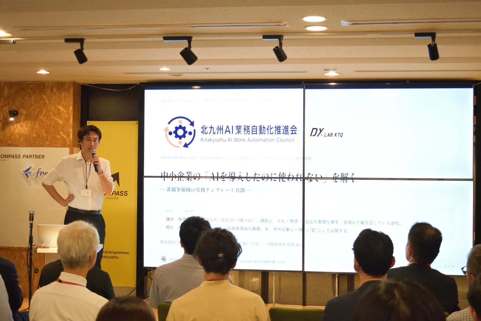 採択者ピッチで登壇する当社代表。スクリーンには北九州AI業務自動化推進会のロゴと「中小企業の『AIを導入したのに使われない』を解く」の表題