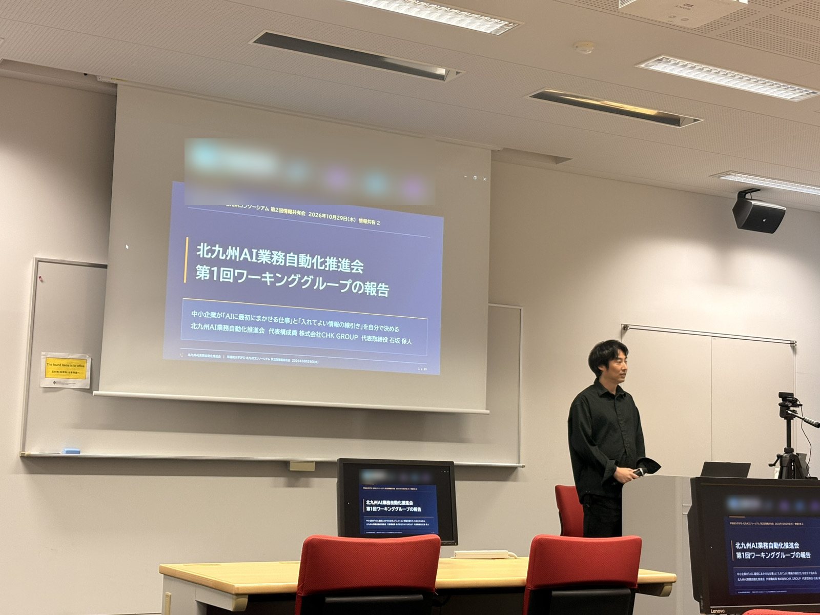 早稲田大学IPS・北九州コンソーシアムの情報共有会で、表紙スライド「北九州AI業務自動化推進会 第1回ワーキンググループの報告」の前に立つ当会の代表