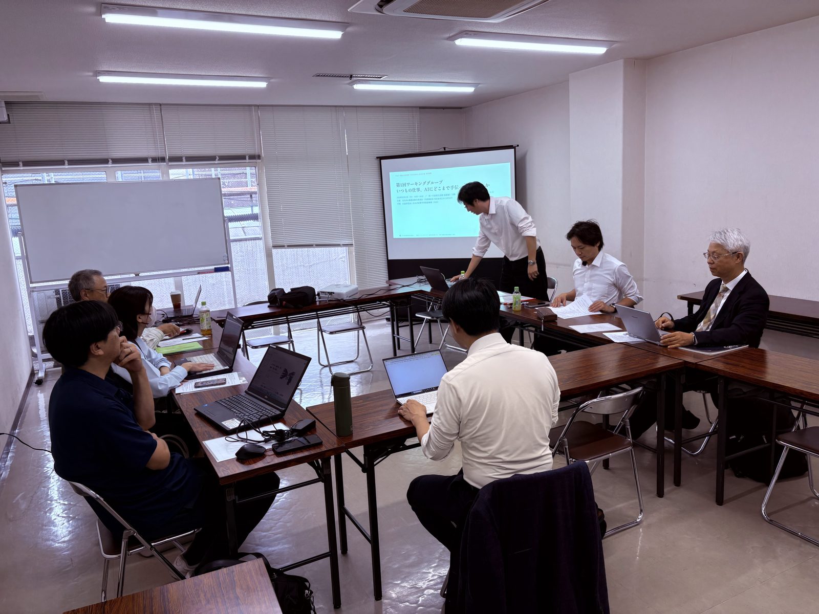 第1回ワーキンググループの会場。コの字に並んだ机に参加者が着席し、前方のスクリーンに表紙スライド